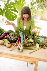 Obraz premium Portrait of a young cheerful sits by the table full of fresh vegetables, fruits and greens indoors with plants on background. Healthy eating and lifestyle concept