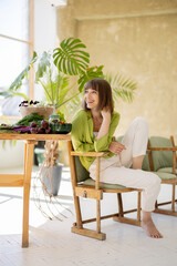 Portrait of a young cheerful woman sits relaxed on chair near the table full of fresh vegan food in the room with green plants on background. Healthy eating and wellness concept