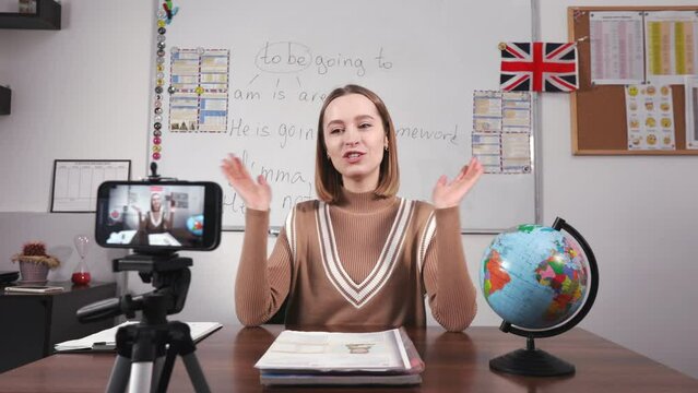 Pleasant english teacher sitting at desk with whiteboard behind and waving on camera while recording video for educational blog. Cheerful woman greeting students using smartphone on tripod at school.