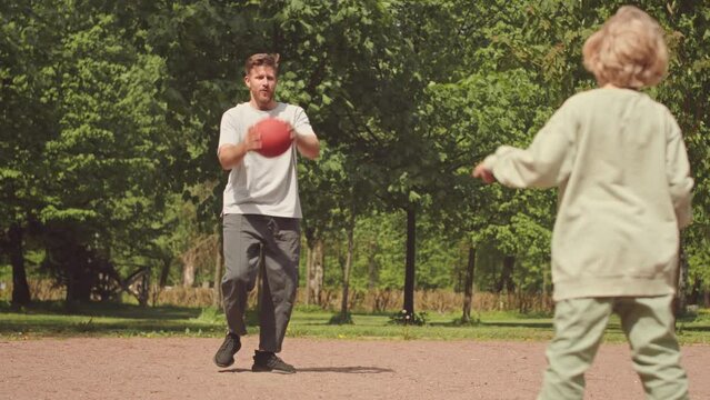 Slowmo of handsome young man and his little son playing with basketball outdoors in park on warm sunny day, throwing it to each other