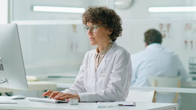 Female doctor with curly brunette hair sitting at desk in clinic and working on computer