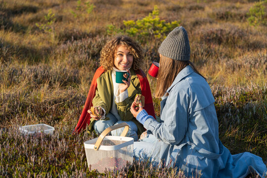 Two trendy girls having cozy autumn picnic in countryside with hot tea, sandwiches enjoying sunny days. Tea break. Happy laughing female friends or sisters travel to woods on weekend together