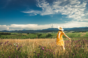 Woman in yellow dress and hat staying at the green meadow in the mountain