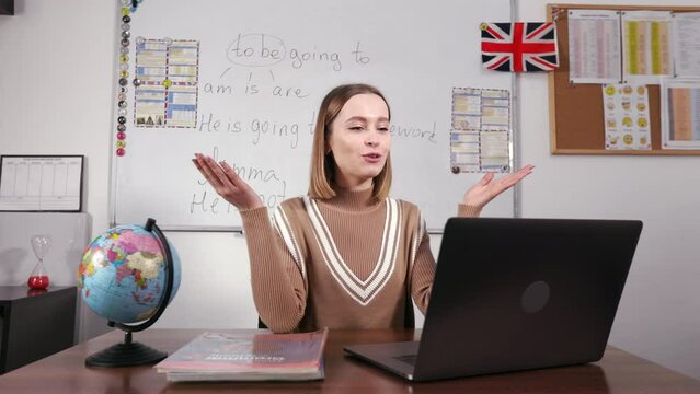 Happy caucasian female teacher sitting at workplace and having video lesson with students. Smiling woman looking at laptop screen, waving with hand and explaining grammar online.