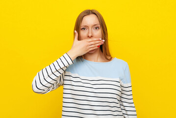 Portrait of young shocked woman with fair-haired gasping, covering mouth and looking startled, standing over yellow background in casual striped sweatshirt