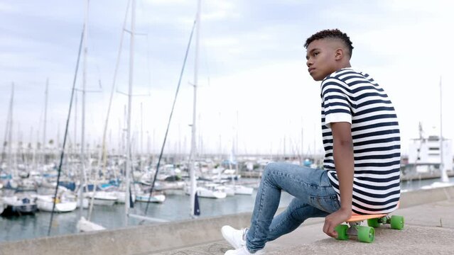 Thoughtful young teenage boy looking away relaxing outdoor sitting on skateboard by sea port. Slow motion high quality 4k footage