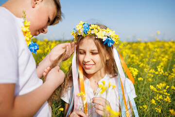 Brother braids ribbons in Ukrainian wreath on head of sister, in rapeseed field under clear sky