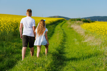 Rear view of children: brother and sister walking far away along path with grass surrounded by yellow fields against sky