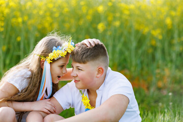 Children enjoying weather: brother and sister sit on squats and talk in field with flowers and grass under sky