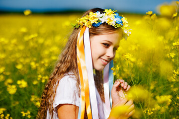 Teenage girl in dress and Ukrainian wreath with lentims on head, in rapeseed field under a clear...