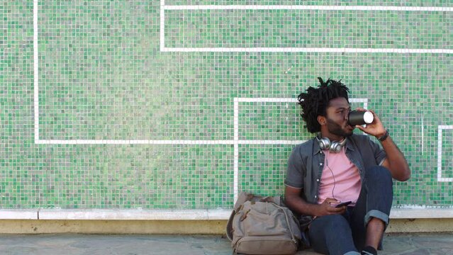 A young trendy black male student with dreadlocks sitting outside against a tiled wall. Stylish African man drinking coffee before answering a phone call and leaving for class in an artistic town