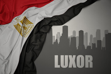 abstract silhouette of the city with text Luxor near waving colorful national flag of egypt on a gray background.