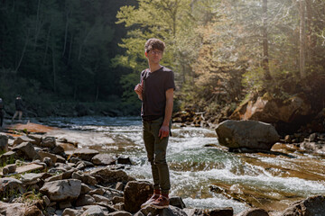 Young male traveler with backpack standing on rocks at river in mountains. Trekking route. 