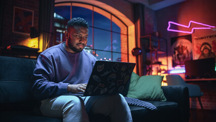 Young Stylish Black Man Working from Home on Laptop Computer with Stickers in Cozy Stylish Loft...
