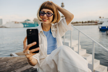 Cheerful young caucasian woman in sunglasses with phone sits by sea. Brown-haired wears casual clothes, enjoys her vacation. Lifestyle concept.