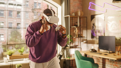Young Male Gamer Having Fun and Playing Virtual Reality Game at Home in His Living Room. Black Man Wearing a Modern Headset and Using Controllers. Stylish Apartment with Big Window. © Gorodenkoff