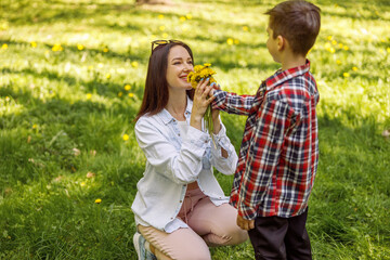 Cheerful young mother smelling flowers from her son. Mom and kid in park with dandelions.