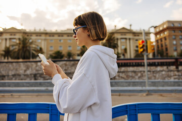 Modern young caucasian woman uses smartphone standing outdoors in city. Brown-haired with bob haircut wears white hoodie and sunglasses. Social media addiction concept