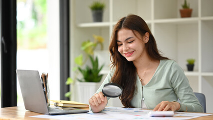 Young female auditor inspecting financial document with a magnifying glass at office desk