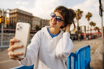 Smiling young caucasian woman communicates by video via phone standing on street. Brown-haired with...