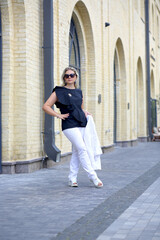 Fototapeta na wymiar Confident business woman in sunglasses in a stylish black blouse and white trousers on a summer morning on a city street. Women's white suit. Fashion concept
