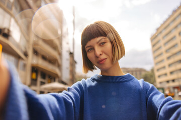Pretty young caucasian girl looking at camera takes selfie outdoors during day. Brown-haired woman...