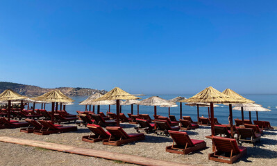 On a rocky beach there are a lot of wooden sunbed  for relaxing. Near them are sun umbrellas with straw tops. In the background is a clear, blue sky and the endless sea.