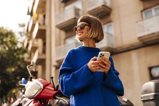 Smiling young caucasian girl with smartphone in her hands, looks away relaxing outdoors. Brown-haired woman wears sunglasses and blue sweater. Happy weekend concept, use