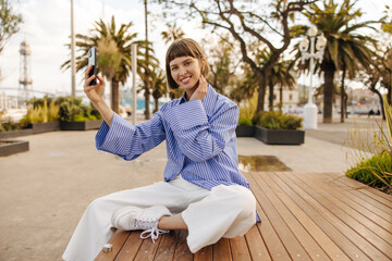 Cheerful young caucasian lady looks at camera, holds phone in front of her sitting on bench during day. Brown-haired woman with bob haircut wears casual clothes. Concept of positive emotions, use