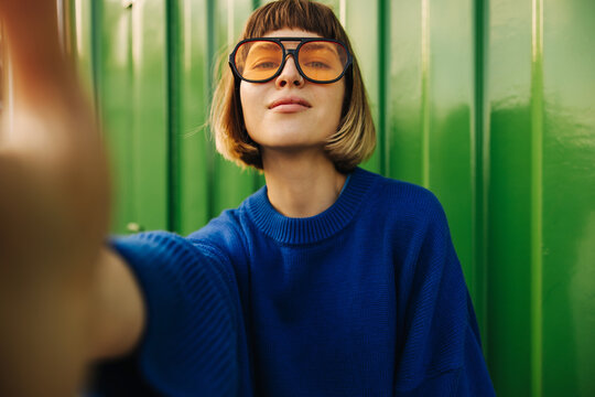 Cute young caucasian girl makes selfie looking at camera on background of green wall street. Brown-haired woman with bob haircut wears blue sweatshirt and sunglasses. Modern lifestyle concept