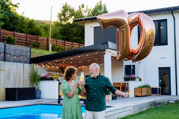 Senior man with number 70 balloons with his wife celebrating birthday and toasting with wine near...