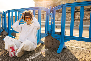 Funny caucasian young girl ruffles her hair looking at camera, sits on pavement during day street....
