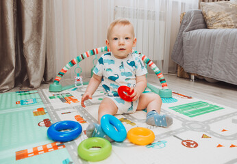 A cute baby is playing with a rainbow toy pyramid while sitting on a play mat in a sunny bedroom. Toys for small children.