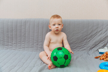 the kid is holding a soccer ball, sitting on the couch barefoot and looking away. Adorable baby is playing at home on the couch. The concept of holidays, weekends and childhood