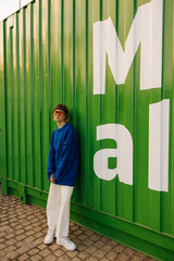 Full length modern young caucasian woman looking at camera standing near green city wall....