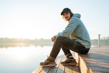 A guy sits on a pier looking into the camera, a curly-haired young man in a cap, a hipster in a...