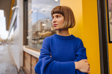 Serious young caucasian girl looks away, arms crossed standing outdoors. Brown-haired woman with bob haircut wears blue sweatshirt in spring. City life concept