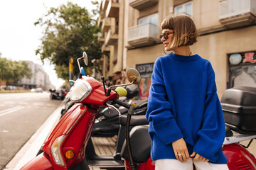 Pretty young caucasian woman in sunglasses sits on moped and looks away at view of city. Brown-haired with a bob haircut wears blue sweater in autumn. City life concept