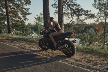Fototapeta na wymiar Young sexy woman biker in bikini sitting on a motorcycle on the side of a forest road