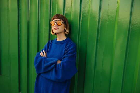 Stylish young caucasian woman posing looking at camera with arms crossed on green wall background. Brown-haired with bob haircut wears sunglasses, blue sweatshirt. Relaxed lifestyle, concept