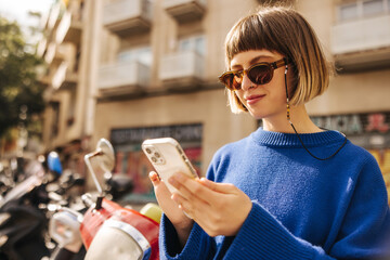 Attractive young caucasian girl is typing message on phone standing outdoors. Brown-haired woman...