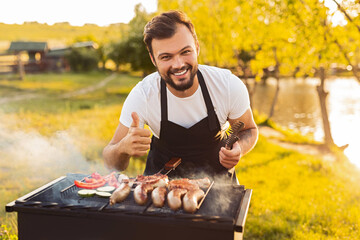 Happy ethnic male chef showing like sign while grilling meat and veggies in park