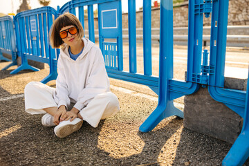 Stylish young caucasian hipster girl looks at camera, sits on pavement along road during day....
