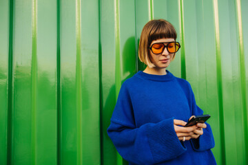 Pleasant young caucasian woman browsing social media on smartphone standing near green wall during...