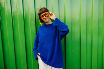 Fashionable young caucasian woman in sunglasses posing against backdrop of green city wall....