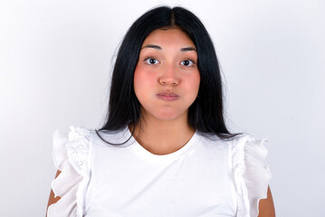 young brunette woman wearing white T-shirt standing against white background puffing cheeks with funny face. Mouth inflated with air, crazy expression. - obrazy, fototapety, plakaty