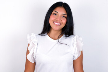 young brunette woman wearing white T-shirt standing against white background with broad smile,...
