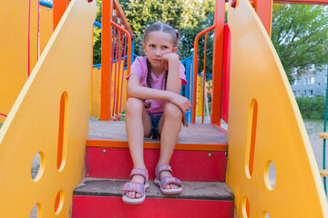 A child girl with blond hair in a pink T-shirt sits offended and sad on the playground. 