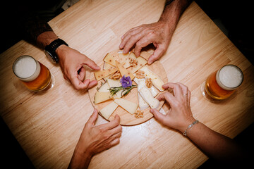 four hands picking up pieces of cheese presented on a wooden board on a table with two beers