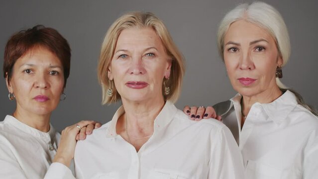 Chest up studio portrait of three confident mature women in white shirts posing for camera standing on gray background